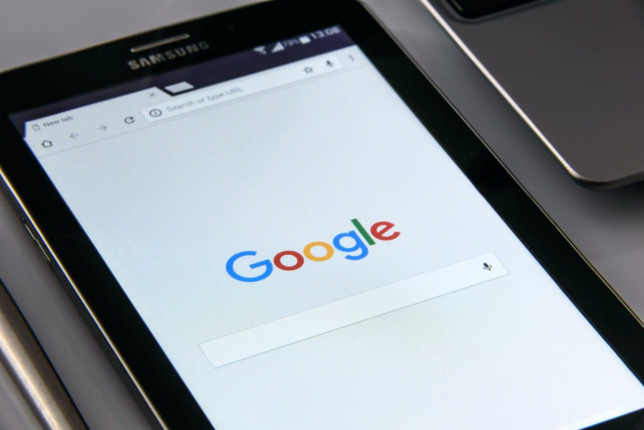 A Samsung tablet displaying the Google homepage with a search bar in the center sits on a white surface beside a silver device, possibly a laptop. Perfect for marketing agencies refining their web design or offering cutting-edge SEO services.