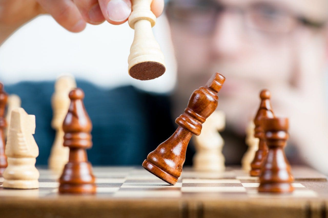 Close-up of a chessboard with a person’s hand strategically moving a white pawn. The black king is tilted, hinting at an imminent checkmate. Other pieces sit in anticipation, as an observer watches intently, much like how a digital marketing company scrutinizes analytics for optimal SEO strategies.