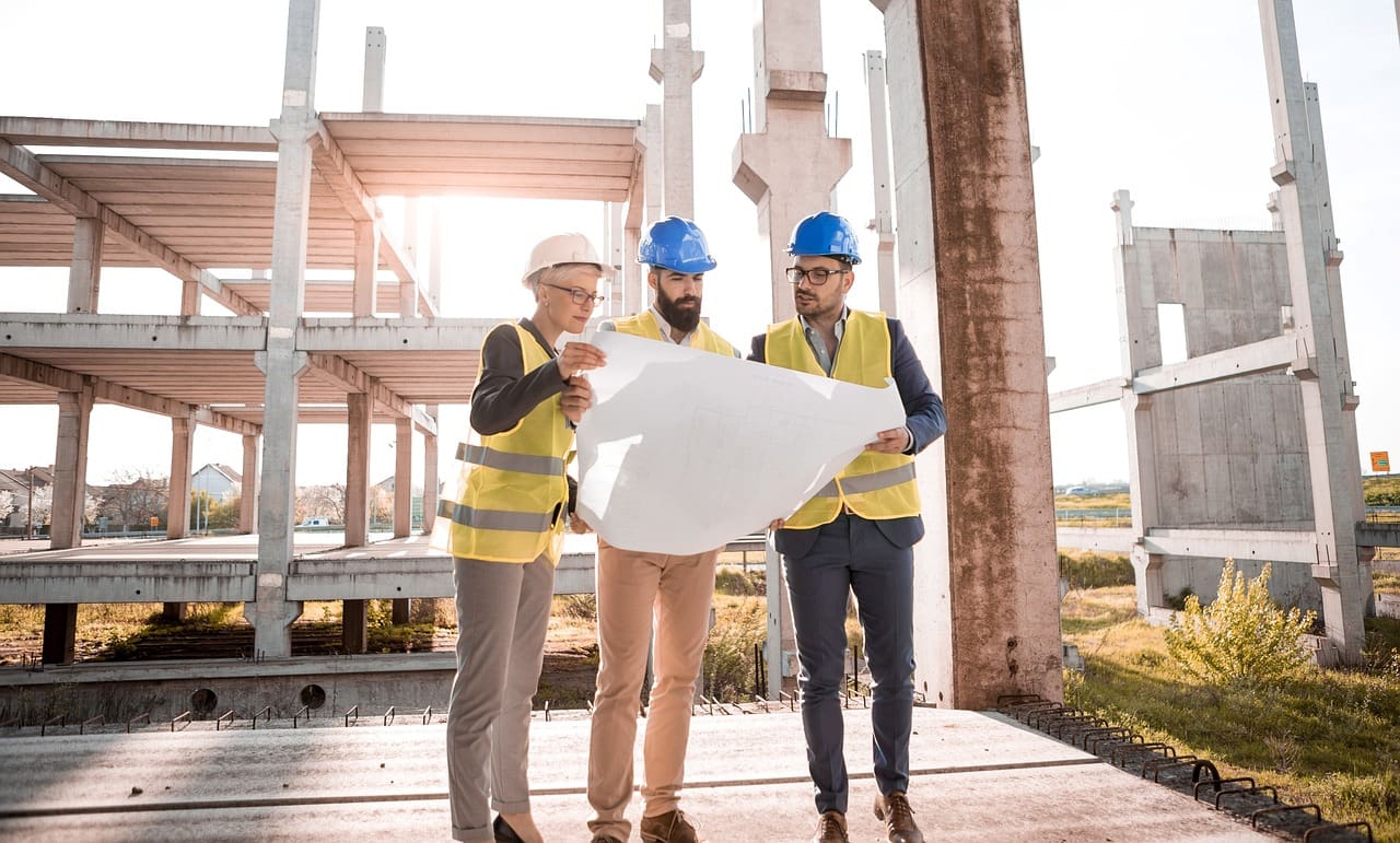 Three construction professionals, two men and one woman, in safety gear discussing blueprints at a building site with concrete structures.
